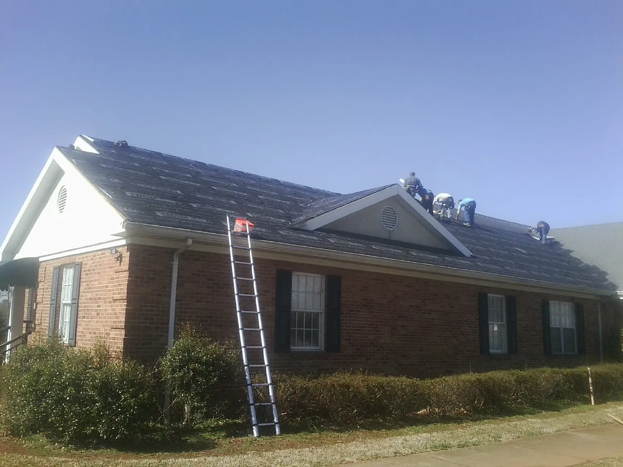 Crew tearing shingles off a brick ranch roof with a ladder set — Emergency Roof Repair work by our crew in Ramey