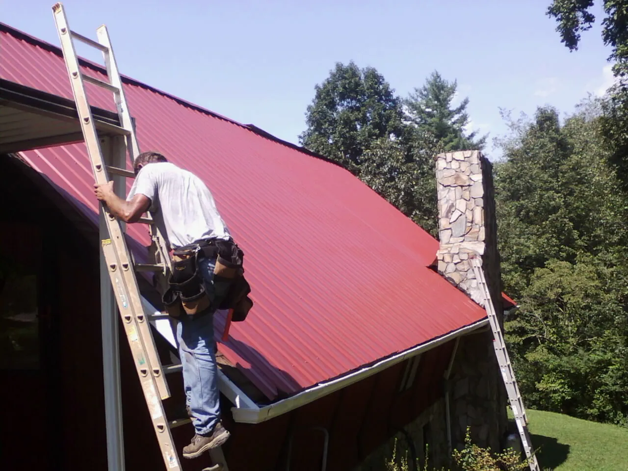 Roofer on a ladder beside a red metal roof and stone chimney — Soffit & Fascia Repair by Roofing Brothers in Ramey